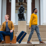 Chris Bird and Kavonte Jones with luggage in front of the Thinker statue on UofL's campus