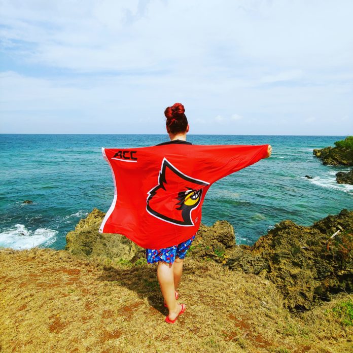 Cheyenne Hill holds a UofL flag overlooking a beach in Trinidad and Tobago.