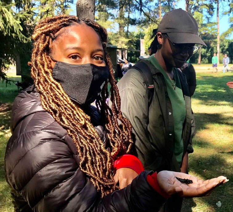 Female student, Kenyetta Johnson, holding a dragonfly in her hand while wearing a black mask on her face.