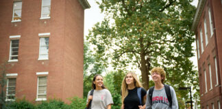 Three students wearing UofL shirts and carrying backpacks walk on a sidewalk on UofL's campus