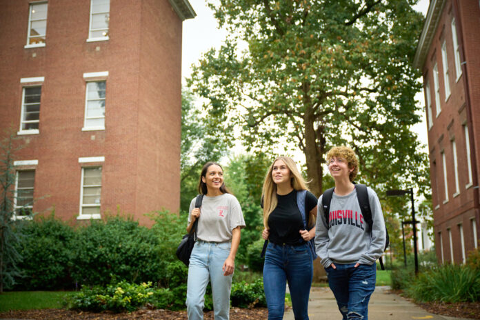 Three students wearing UofL shirts and carrying backpacks walk on a sidewalk on UofL's campus