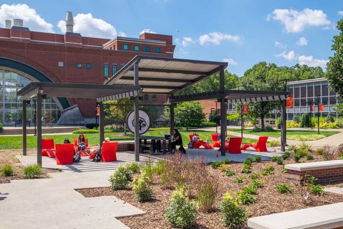 students at tables, chairs, underneath a pergola on a lawn