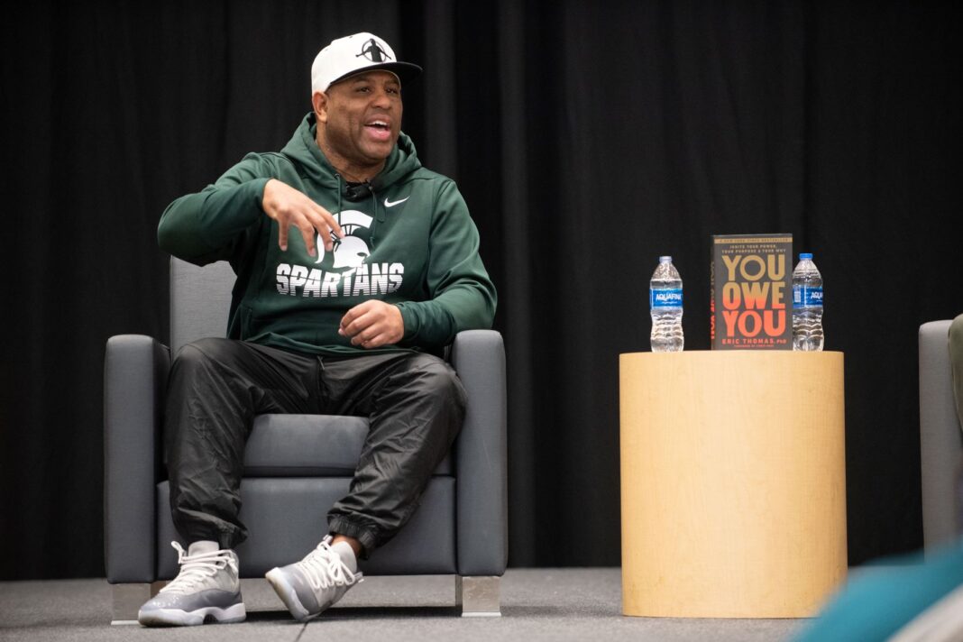 Eric Thomas speaking, seating on stage, wearing ball cap and Michigan State Spartans hoodie