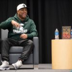 Eric Thomas speaking, seating on stage, wearing ball cap and Michigan State Spartans hoodie