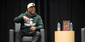 Eric Thomas speaking, seating on stage, wearing ball cap and Michigan State Spartans hoodie