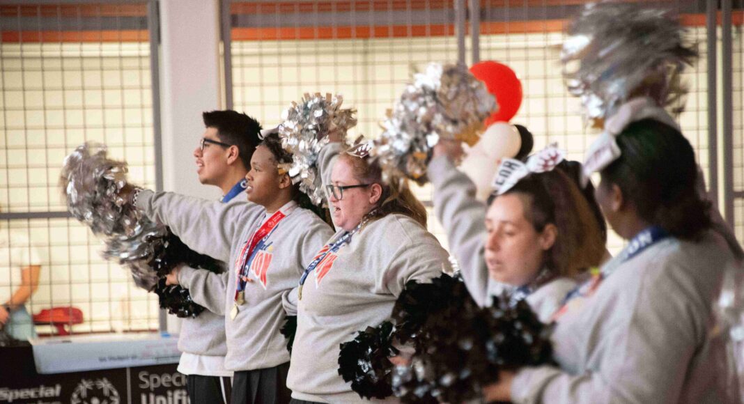 Five cheerleaders in a row, facing left, holding pom-poms. They are wearing grey sweatshirts and skirts or pants. They are standing in an indoor space with a basketball hoop and a banner with "Special Olympics Unified Champion Schools" visible in the background.