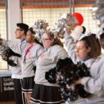 Five cheerleaders in a row, facing left, holding pom-poms. They are wearing grey sweatshirts and skirts or pants. They are standing in an indoor space with a basketball hoop and a banner with "Special Olympics Unified Champion Schools" visible in the background.