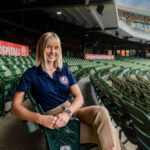a woman in professional clothing sits at a baseball stadium