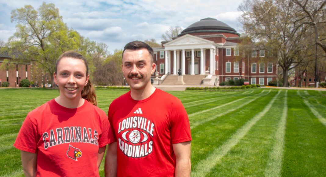 Two people wearing UofL tshirts smile in a grassy lawn.