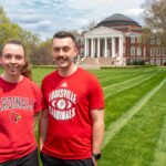 Two people wearing UofL tshirts smile in a grassy lawn.