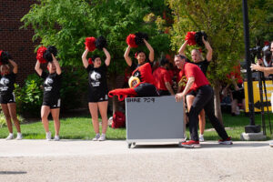 Man pushing the UofL mascot, Louie in a moving cart. 