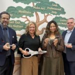 Two men and two women with oversized scissors and ceremonial keys standing in front of a wall mural of a tree with mental health tips