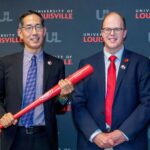 Two men smiling in business suits, one holds a commemorative bat
