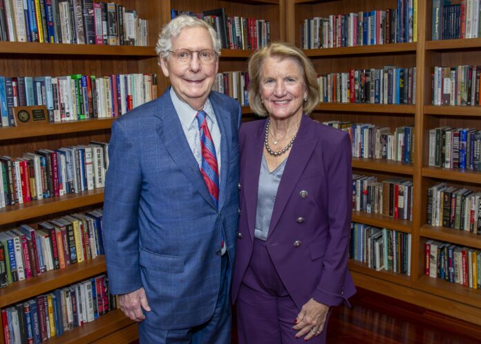 Man and woman stand in a library with shelves of books behind them.