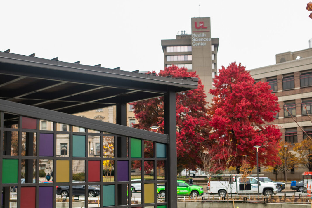 Pergola with colorful panels, red trees and UofL tower in background.