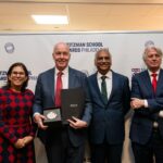 Four people in business attire, one holding a folder and a plaque