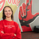 A smiling Carly Lasher crosses her arms in front of a UofL mural.