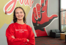A smiling Carly Lasher crosses her arms in front of a UofL mural.