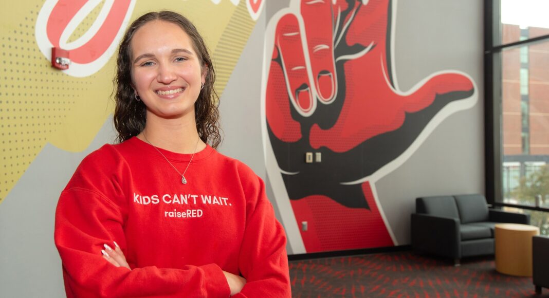 A smiling Carly Lasher crosses her arms in front of a UofL mural.