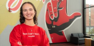 A smiling Carly Lasher crosses her arms in front of a UofL mural.
