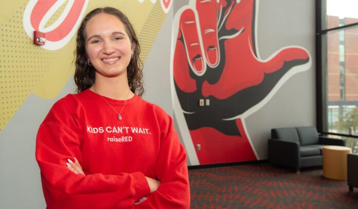 A smiling Carly Lasher crosses her arms in front of a UofL mural.