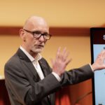 Man discusses information on slides on a screen behind him.