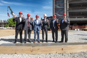 A group of individuals wearing hard hats and holding up the 'L' sign at a construction site. 