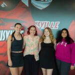 Abby Breeze, Kayla Martinez, Sam Mazzaro and Leena Adapa smile in front of a mural of Louisville athletic jerseys