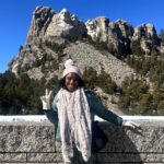 Young woman standing in front of stone wall with Mt. Rushmore monument in background.