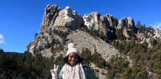 Young woman standing in front of stone wall with Mt. Rushmore monument in background.