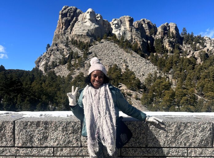 Young woman standing in front of stone wall with Mt. Rushmore monument in background.