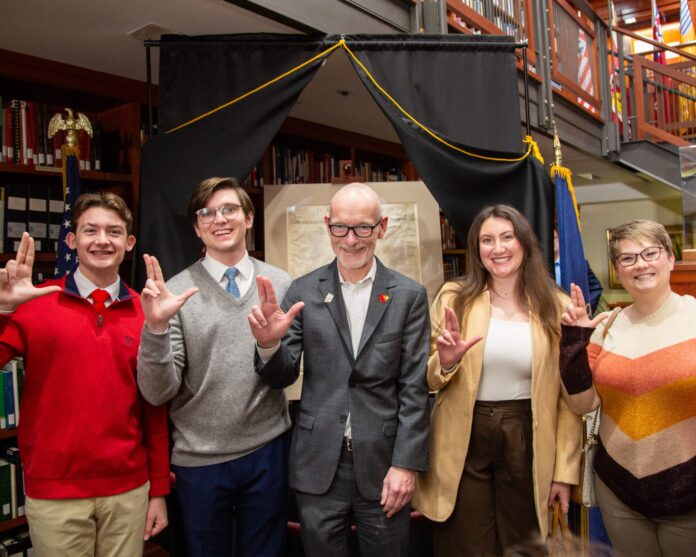 Individuals hold up the L sign with a copy of the Declaration of Independence on display in the background.