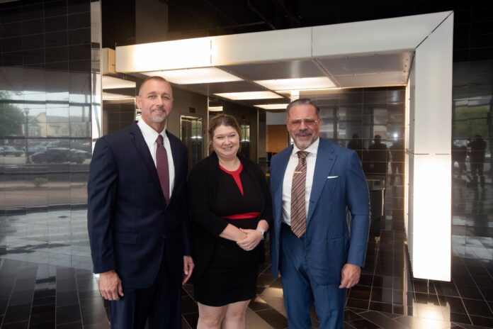 Two men in business suits and a woman in a black dress with red trim.