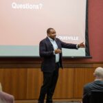 Darrell Clark speaks with his hands animated in front of a screen that states "Questions?"