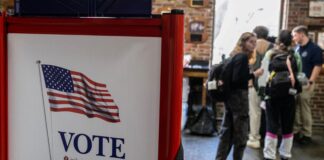 A red, white and blue voting booth with an American flag and the word “VOTE,” with students talking to a poll worker in the background.