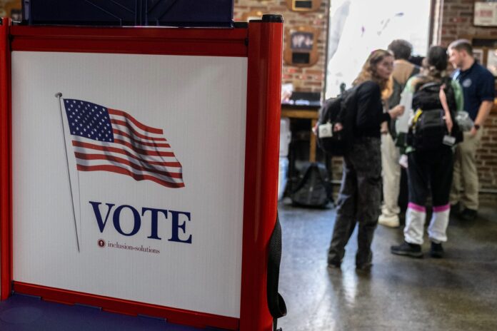 A red, white and blue voting booth with an American flag and the word “VOTE,” with students talking to a poll worker in the background.