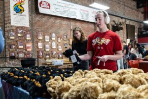 A student in headphones and a red T-shirt holds a black stuffed bear above a table covered in black and tan bears and brown teddy bears, inside the brick-walled Red Barn.