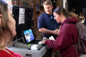 A student in a maroon hoodie feeds a paper ballot into a voting machine labeled Jefferson County Clerk’s Office while a staff member in a navy polo guides her.